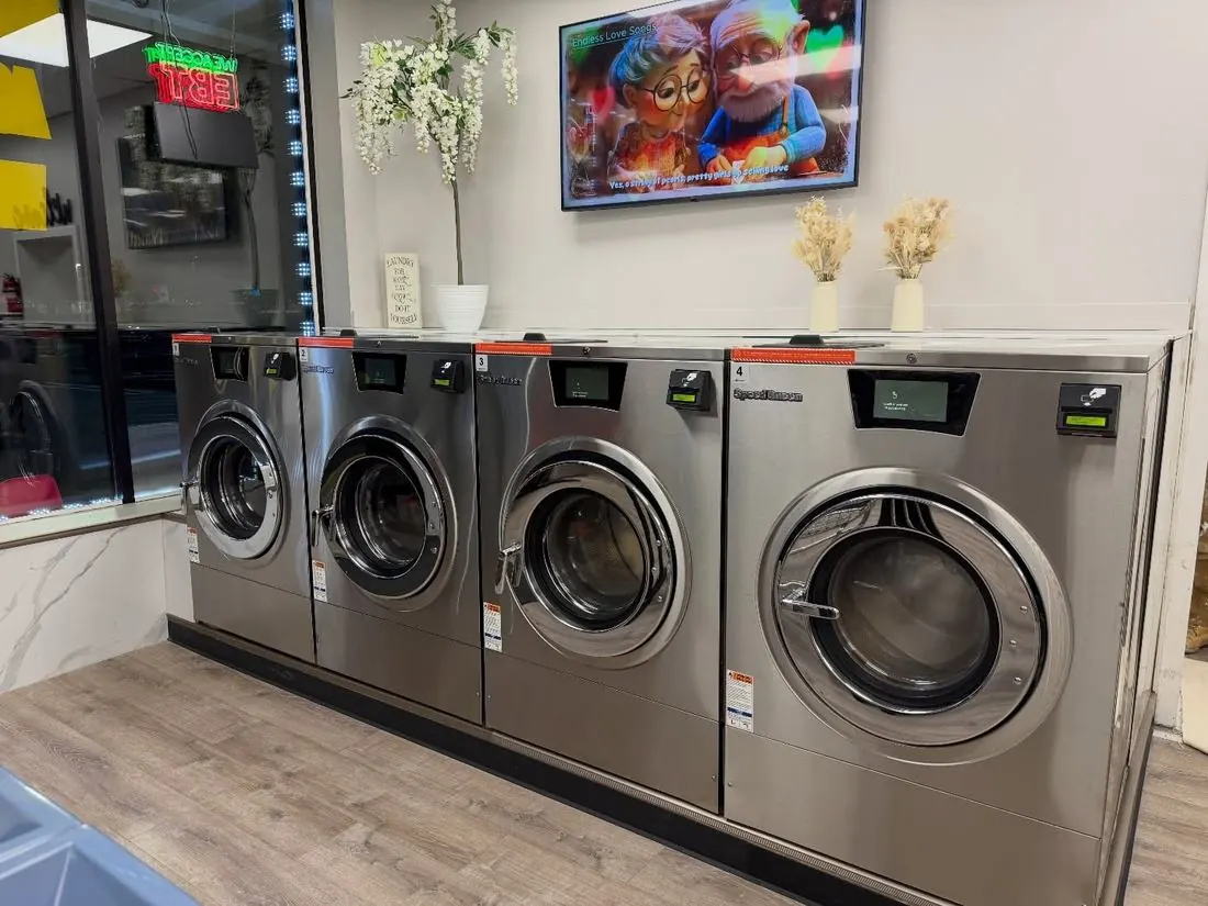 Stainless Speed Queen washers beneath a wall-mounted TV at Newburgh Express Laundry